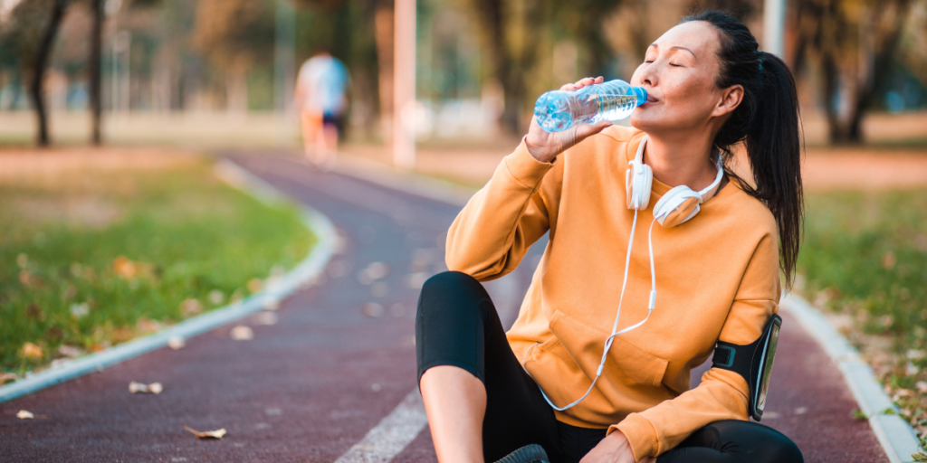 A woman sitting on the ground after a good run, drinking water.