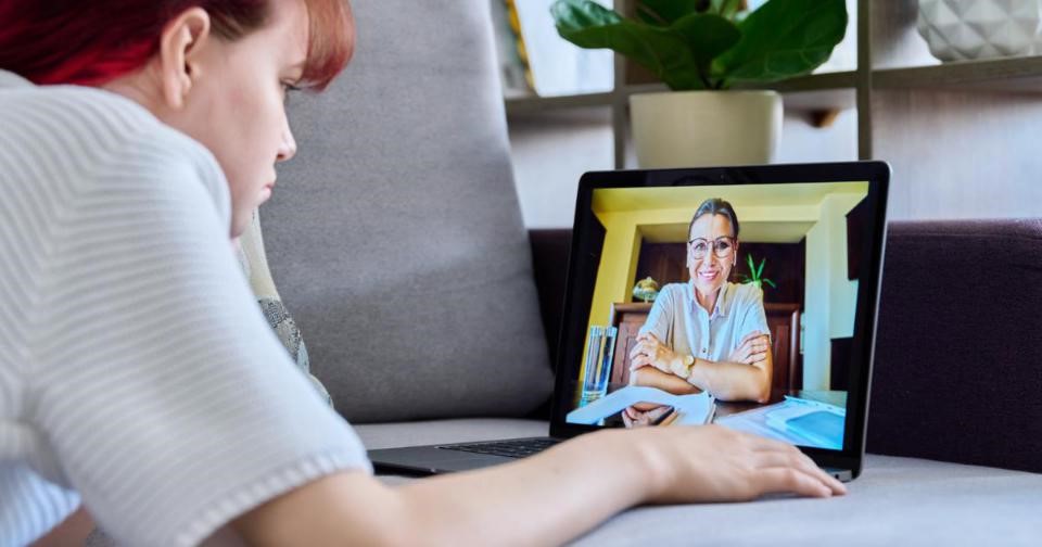 A woman sitting on a couch with a laptop, another woman on the screen. Image depicts virtual therapy session.
