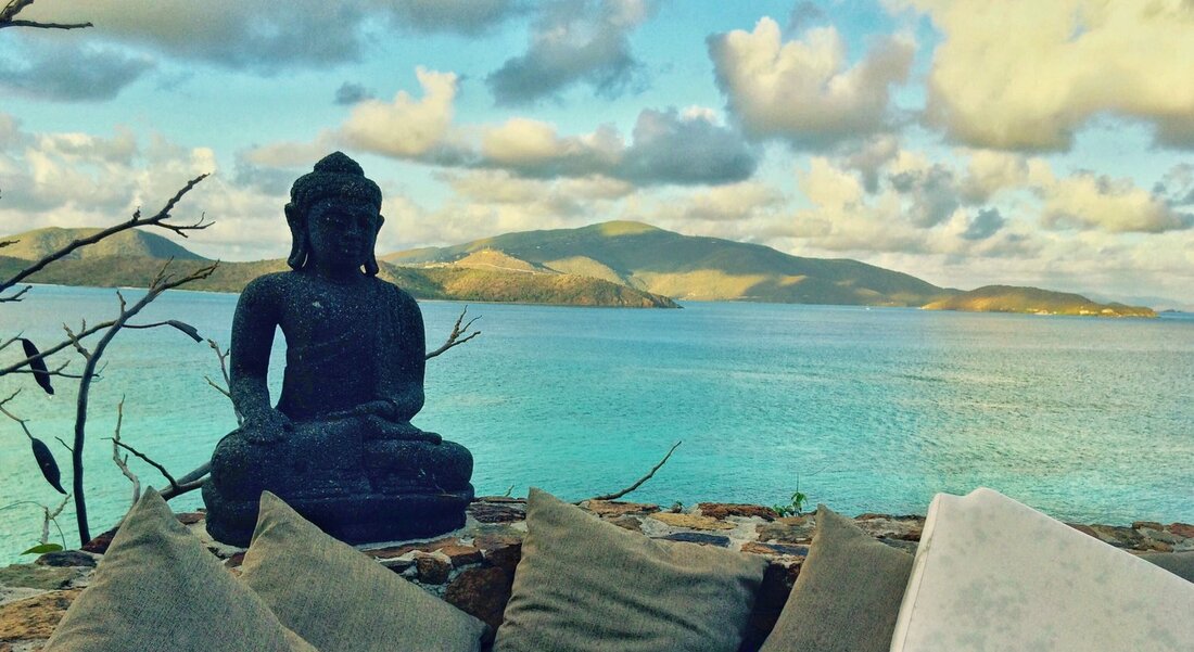 Serene coastal view with Buddha statue and pillows overlooking turquoise ocean and distant green islands under a partly cloudy sky